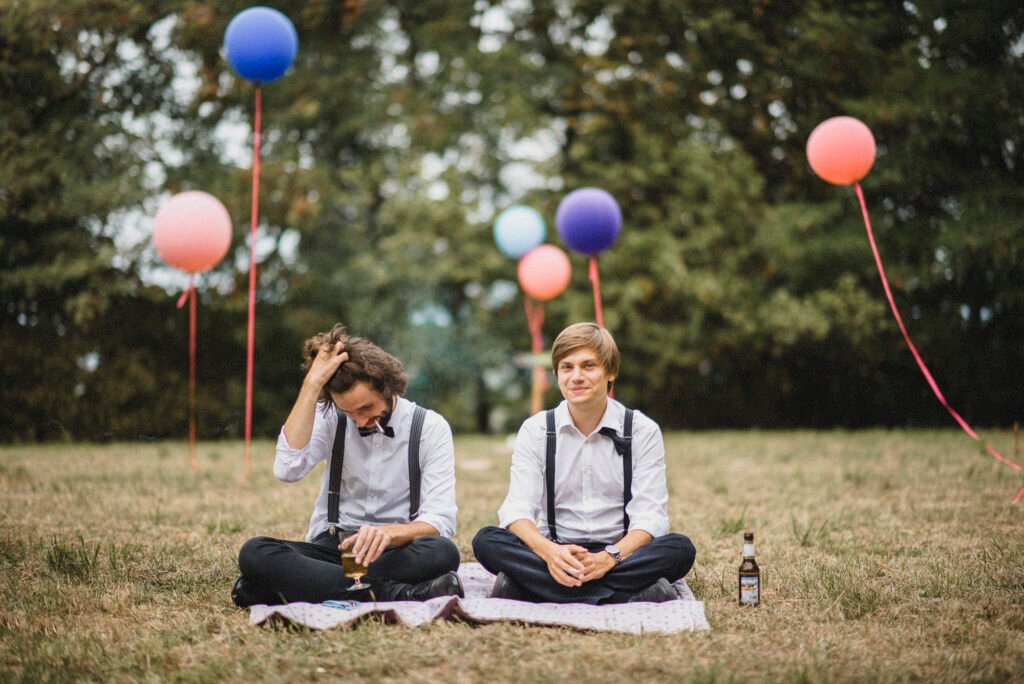 Two grooms wearing suspenders sitting casually on a picnic blanket during their outdoor wedding near Cologne, Germany – authentic wedding photography by a wedding photographer Cologne